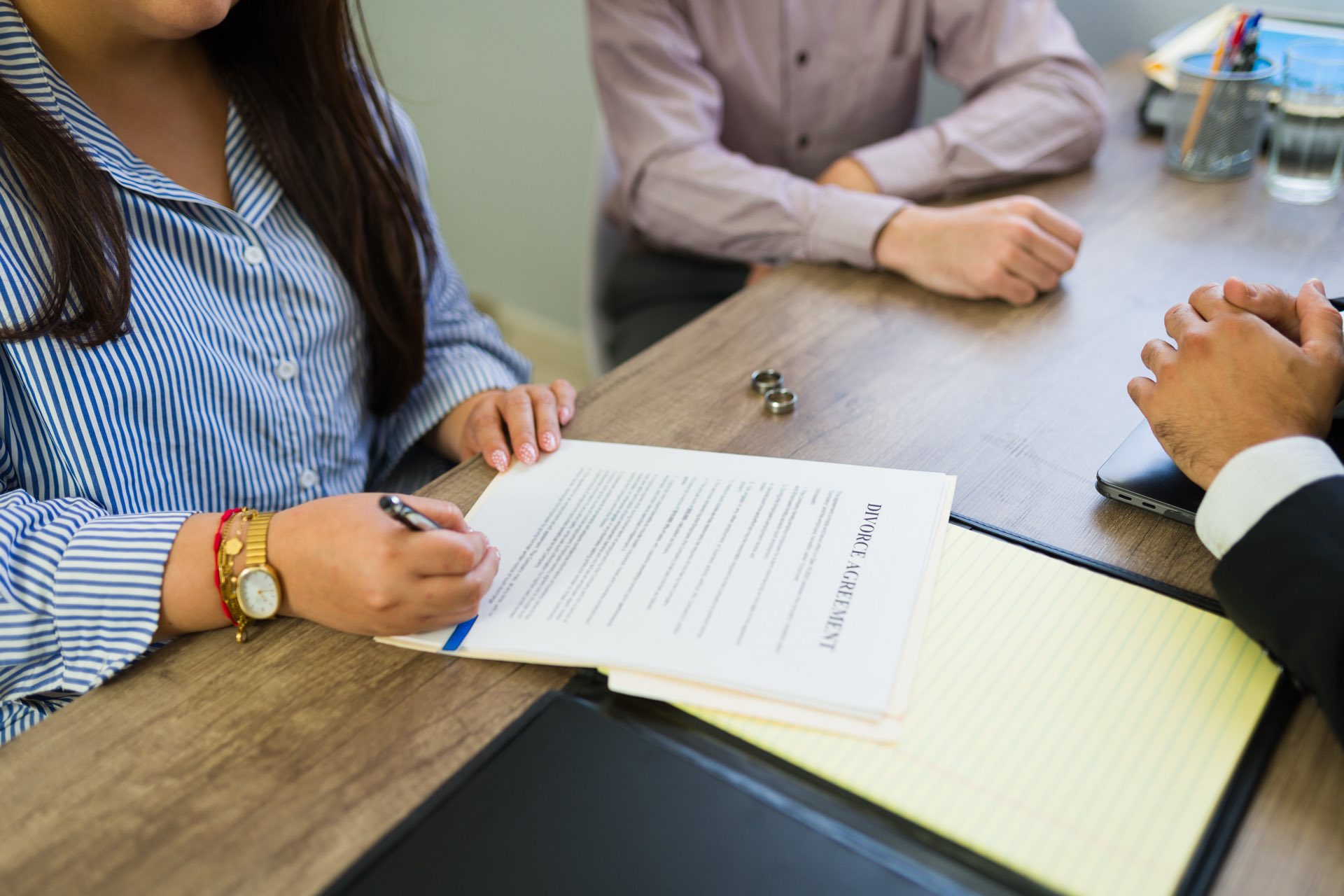 Close up of three people sitting at a table signing paperwork with a set of wedding rings between two of the people